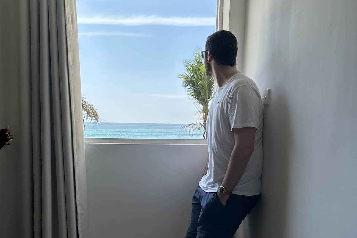 A person leaning against a wall by a window overlooking a beach.