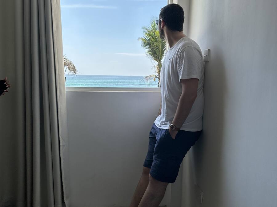 Una persona apoyada en una pared junto a una ventana con vista a la playa.