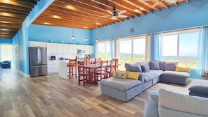 A spacious living area with a large gray sectional sofa, wooden dining table, and chairs. A kitchen with white cabinets and a refrigerator is visible in the background. - 3bd 2 bth House. POOL! HOTTUB! Breathtaking Ocean Views! 2-min drive to beach! (Santos Guardiola)