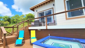 A wooden deck with two lounge chairs, a hot tub, and a staircase leading to a balcony with a view of trees and a clear sky. - 3bd 2 bth House. POOL! HOTTUB! Breathtaking Ocean Views! 2-min drive to beach! (Santos Guardiola)