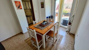 Comedor con una mesa de madera y sillas blancas, una cocina al fondo y vistas al exterior por una puerta.