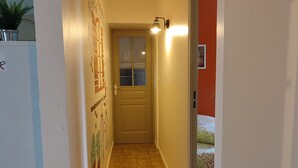 A narrow hallway with a tiled floor, a door with a window, and a wall-mounted light fixture. - Le Gîte du Tilleul (Pernes-les-Fontaines)
