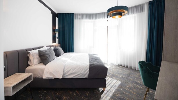 A modern hotel room with a large bed, a wooden nightstand, a green armchair, and a large window with sheer curtains. - Opera Hotel  (Timisoara)