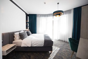 A modern hotel room with a large bed, a wooden nightstand, a green armchair, and a large window with sheer curtains.