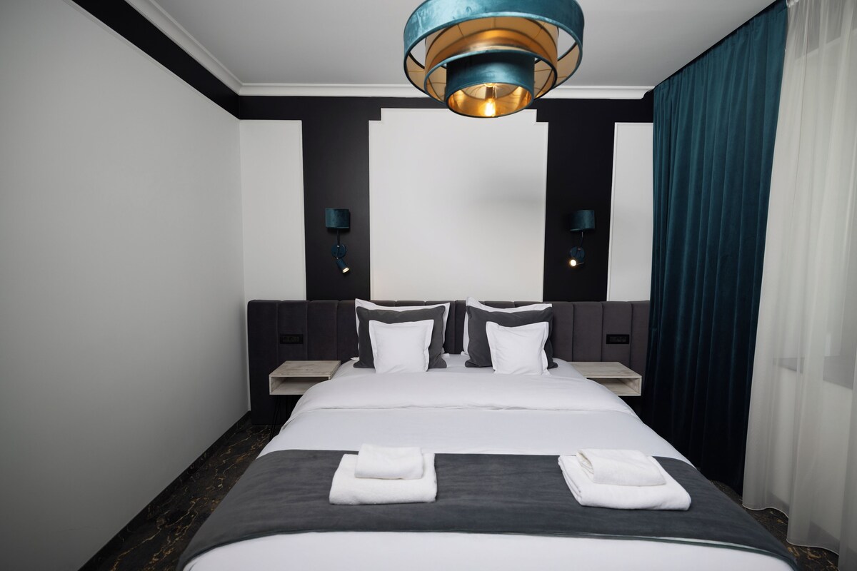 Habitación de hotel con una cama grande, dos mesitas de noche, una lámpara moderna y vistas a la ciudad.