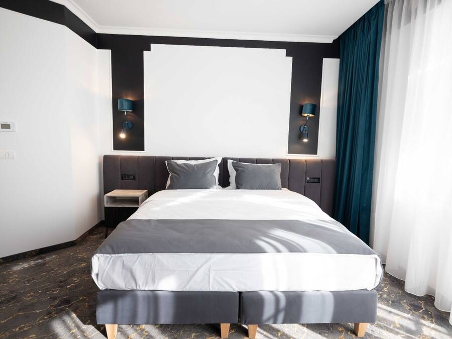 A modern hotel room with a large bed, two bedside tables, a wall-mounted TV, and a dark-colored curtain.