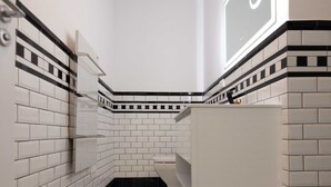 A modern bathroom with white subway tiles, black diamond-patterned flooring, and a large mirror with a light fixture above it. - Opera Hotel  (Timisoara)
