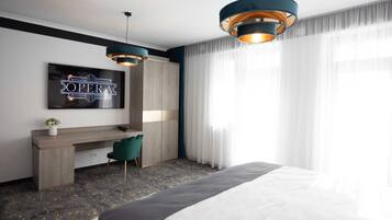 A modern hotel room with a bed, a desk, two pendant lights, a TV on the wall, and sheer curtains.