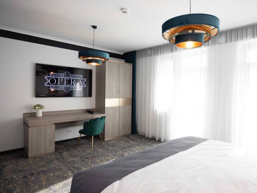 A modern hotel room with a bed, a desk, two pendant lights, a TV on the wall, and sheer curtains.