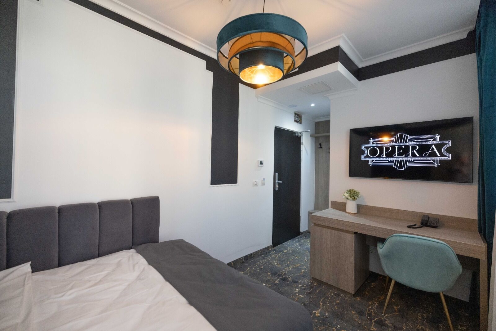 A modern hotel room with a bed, a desk, a chair, a television displaying 'The Opera', and a unique ceiling light fixture.