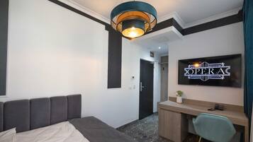 A modern hotel room with a bed, a desk, a chair, a television displaying 'The Opera', and a unique ceiling light fixture.