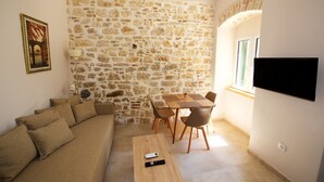 A living room with a stone wall, a wooden dining table with chairs, a sofa with cushions, a remote control on the table, and a TV mounted on the wall. - Galielo Boutique Suite by Corfuescapes  (Corfu)