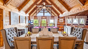 Interior de una cabaña de madera con comedor, mesa de billar y vistas al exterior.