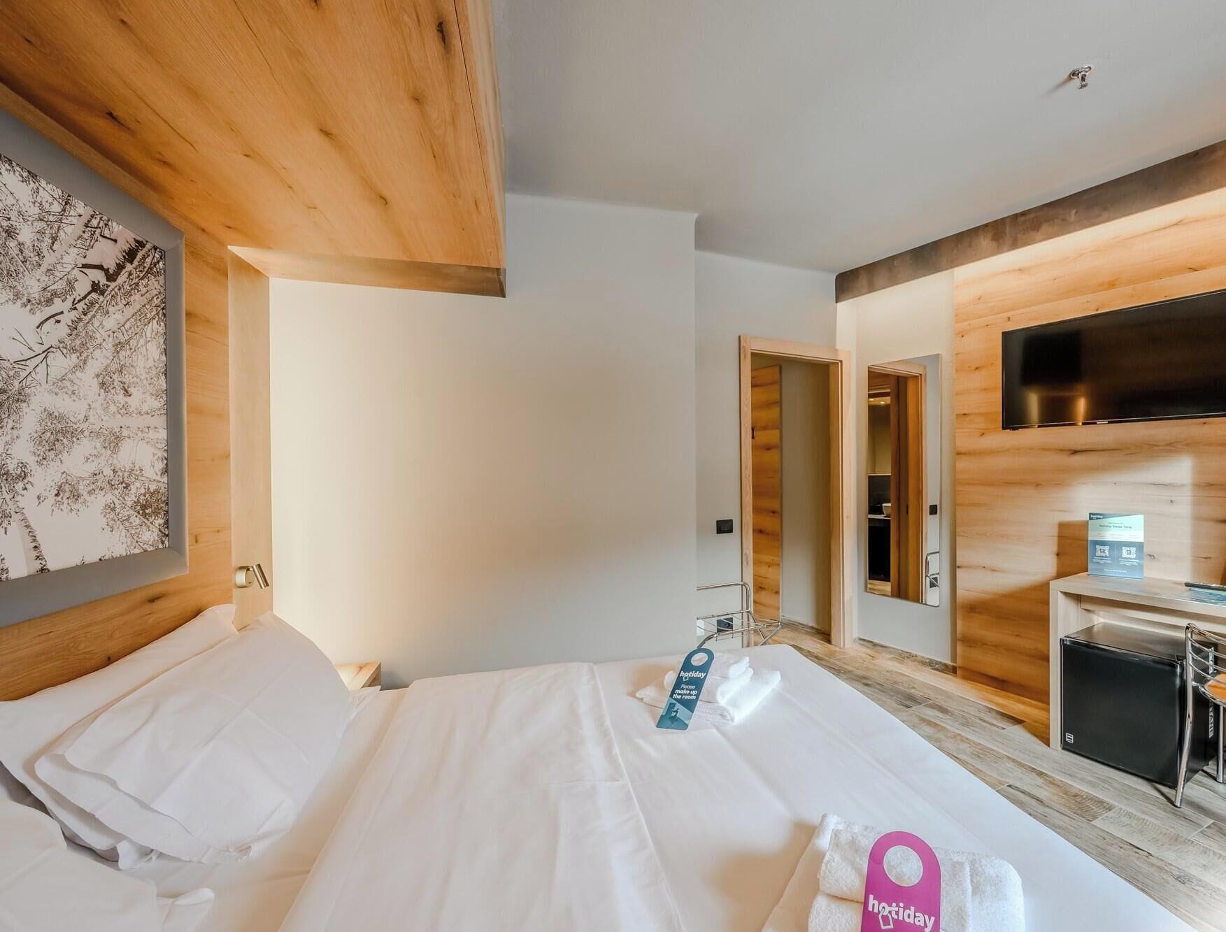 A hotel room with a bed, a wooden headboard, a flat-screen TV, a desk with a computer, and a mirror.