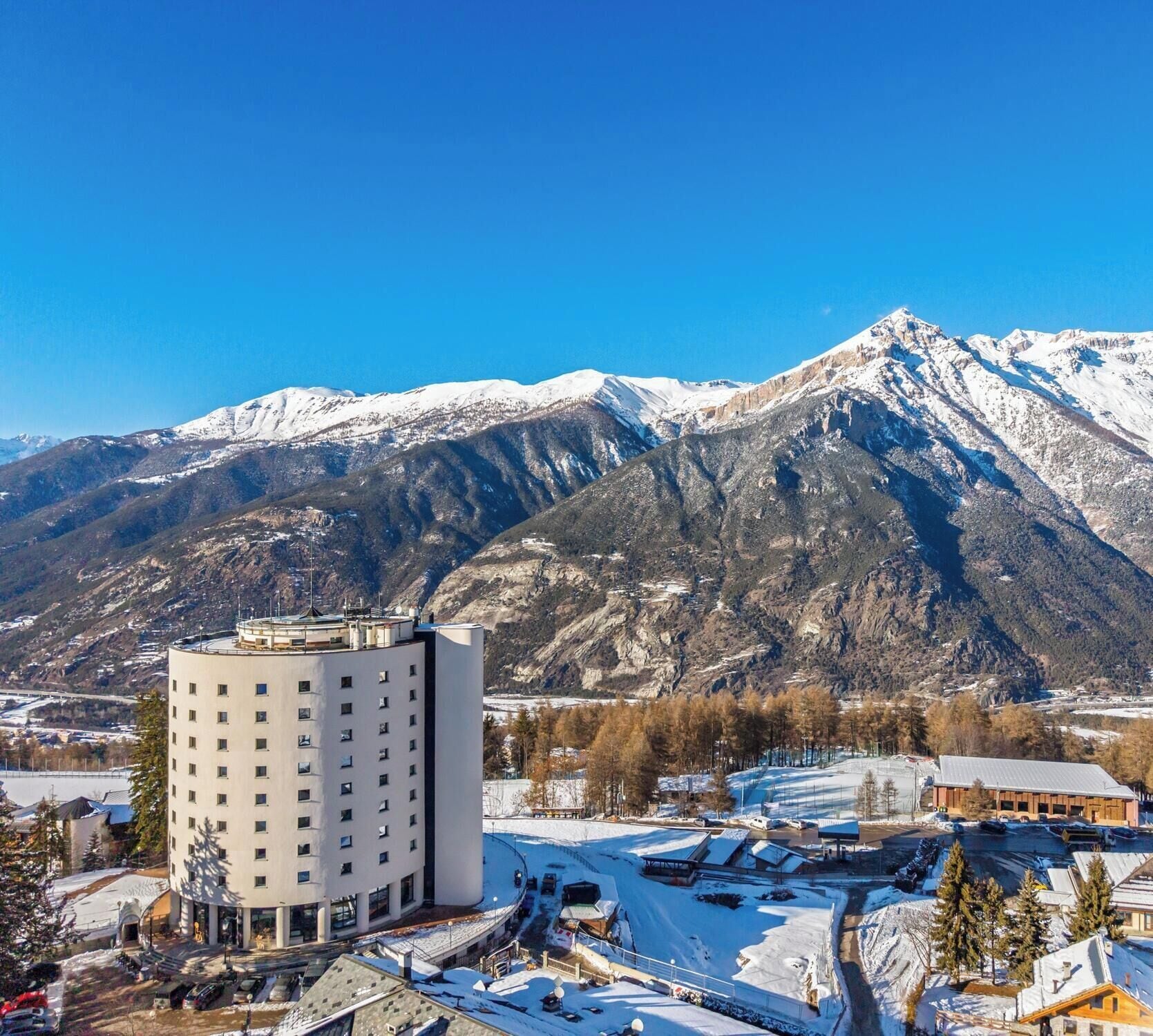 A hotel with a modern design, a parking area, and a view of snow-capped mountains.