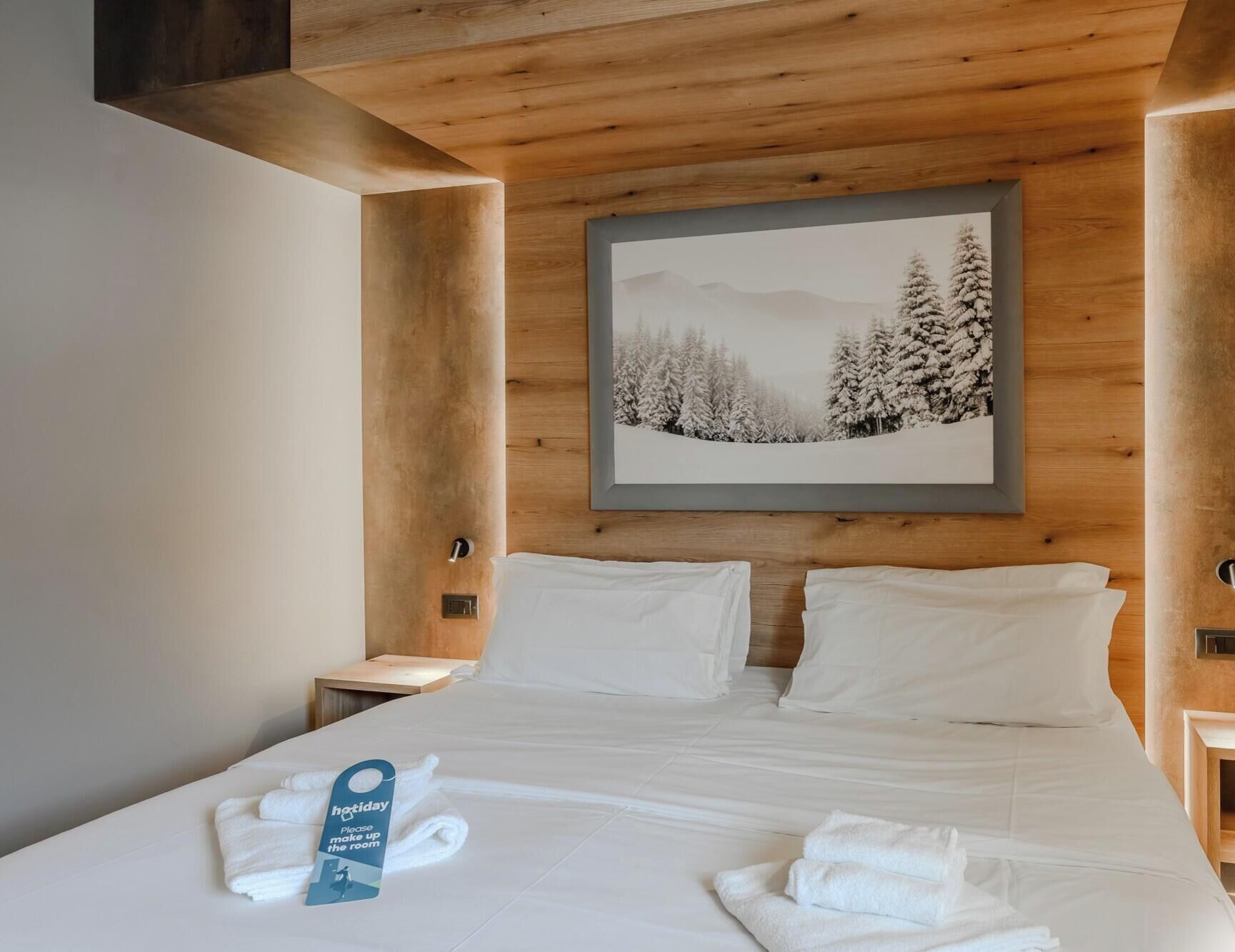 A hotel room with a bed, white linens, a framed picture of a snowy landscape, and a towel with a tag.