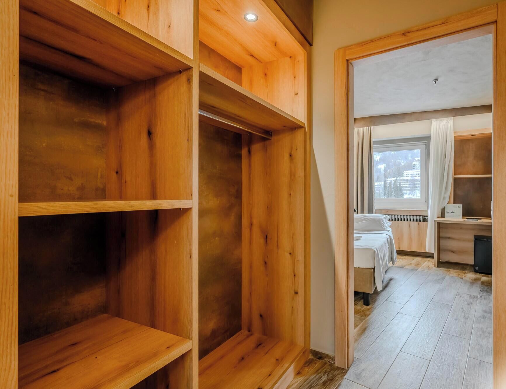 A hotel room with wooden shelving units, a bed, a desk, and a window with curtains.