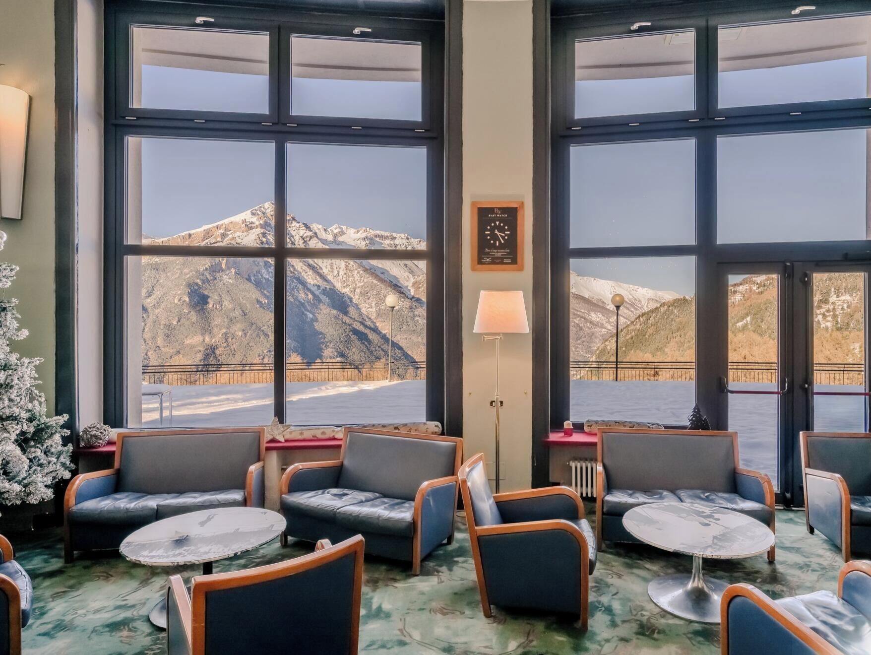 Une pièce dotée de grandes fenêtres donnant sur les montagnes, de plusieurs coins salon avec des tables et d’une horloge au mur.