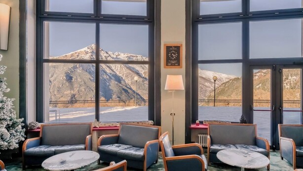 Habitación de hotel con amplios ventanales que ofrecen vistas a las montañas, una zona de estar acogedora con sofás y mesas, y un reloj en la pared.