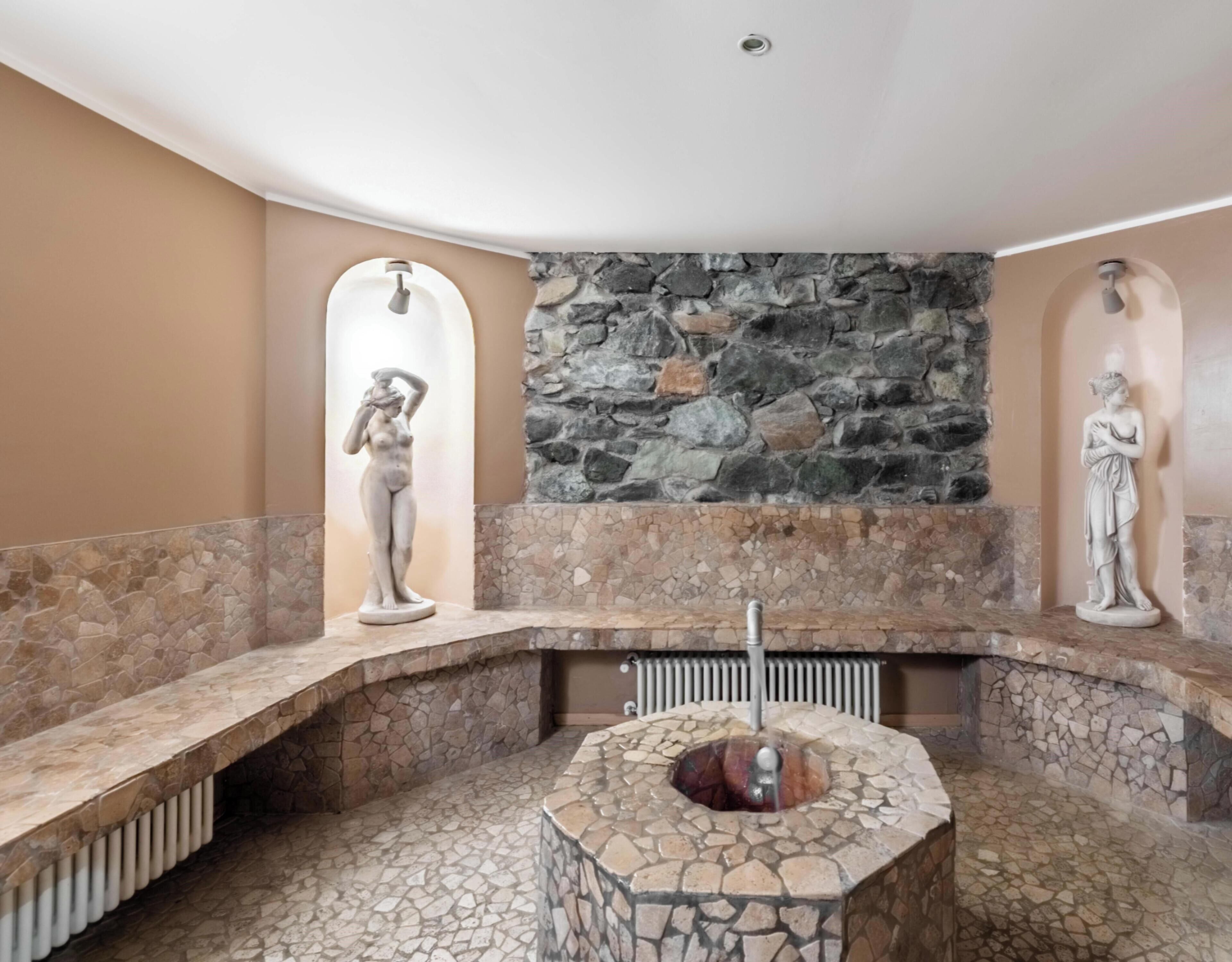 A stone-tiled bathroom with a central circular feature, possibly a sink or basin, flanked by two stone walls with statues.