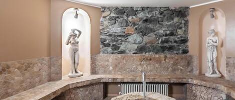 A stone-tiled bathroom with a central circular feature, possibly a sink or basin, flanked by two stone walls with statues.