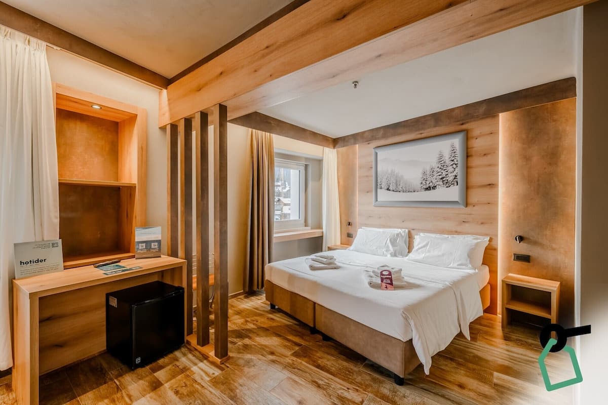 A modern hotel room with a wooden floor, a bed with white linens, a desk with a computer, and a window with curtains.