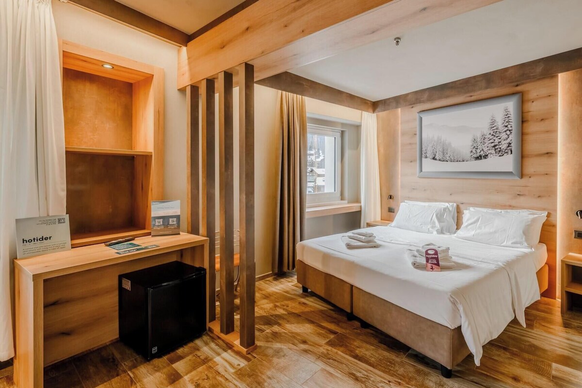 Una habitación de hotel moderna con suelo de madera, una cama con ropa blanca, un escritorio con computadora y una ventana con cortinas.