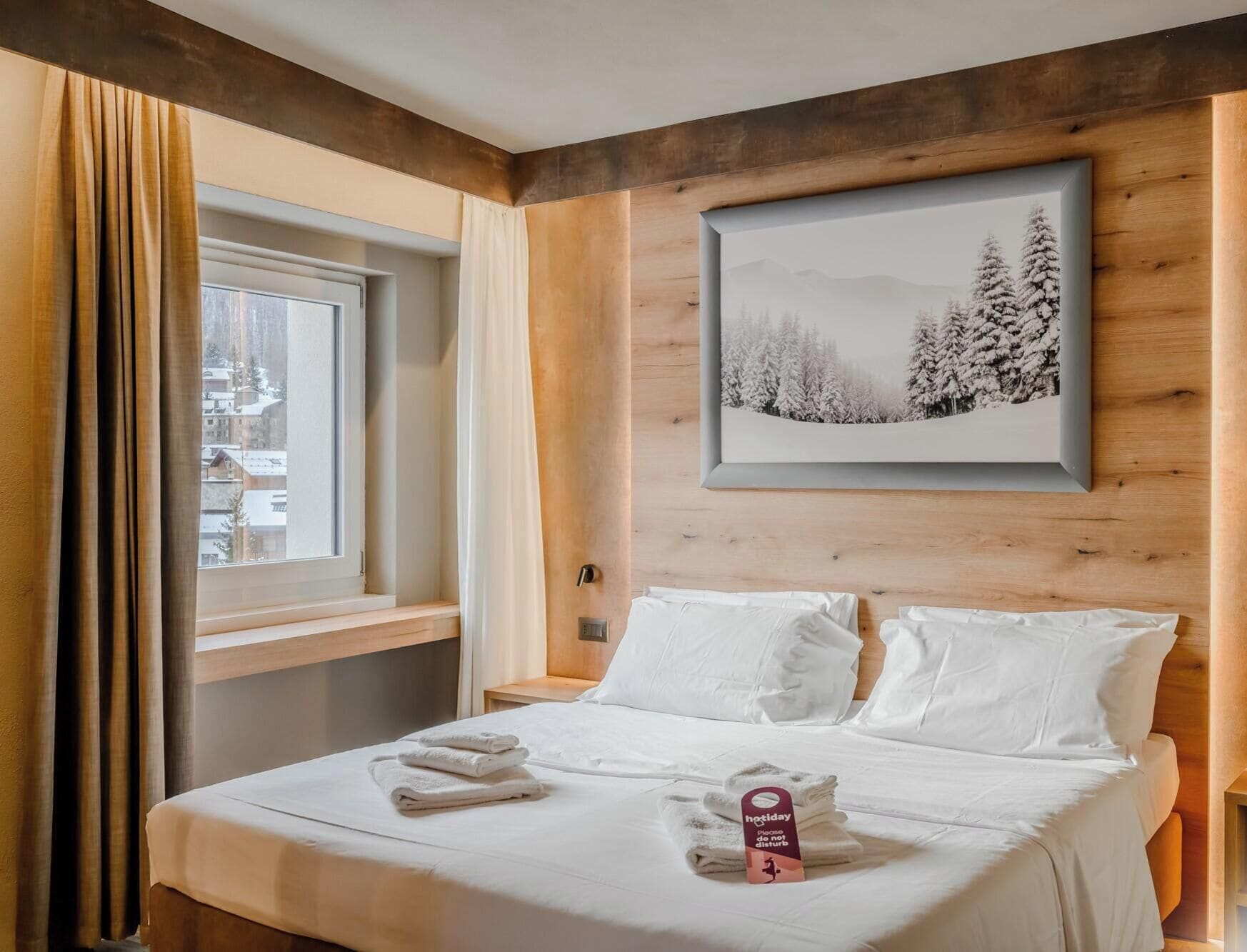 A hotel room with a bed, a window with curtains, a framed picture on the wall, and a tag on the bed.