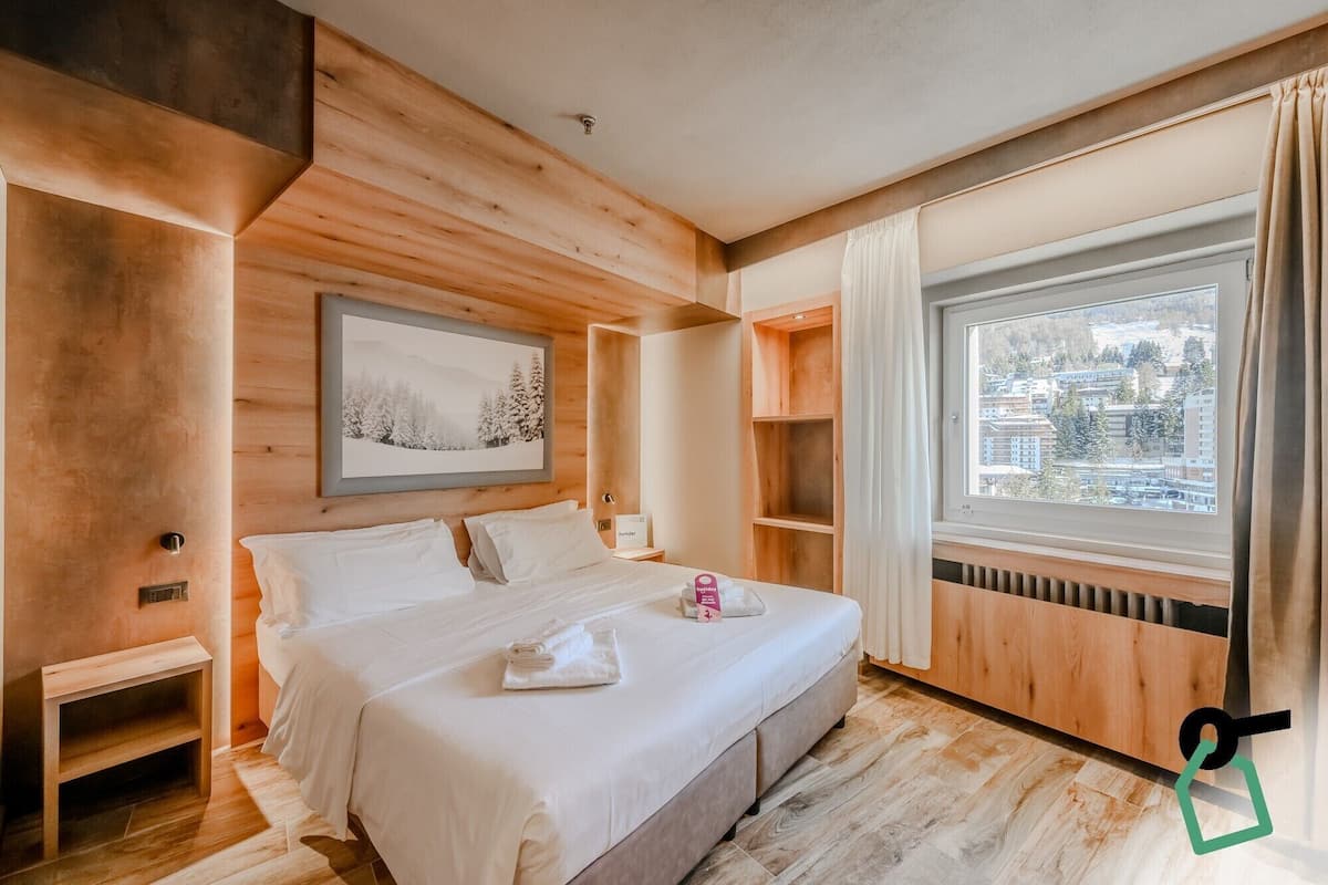 A modern hotel room with a wooden interior, a large bed, a window with a city view, and a framed picture on the wall.