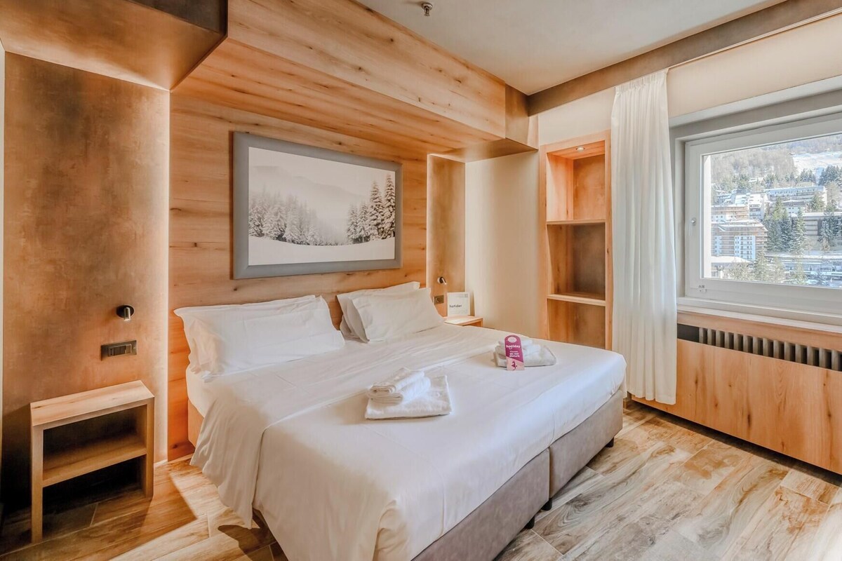 Habitación de hotel moderna con interior de madera, una cama grande, una ventana con vistas a la ciudad y un cuadro en la pared.