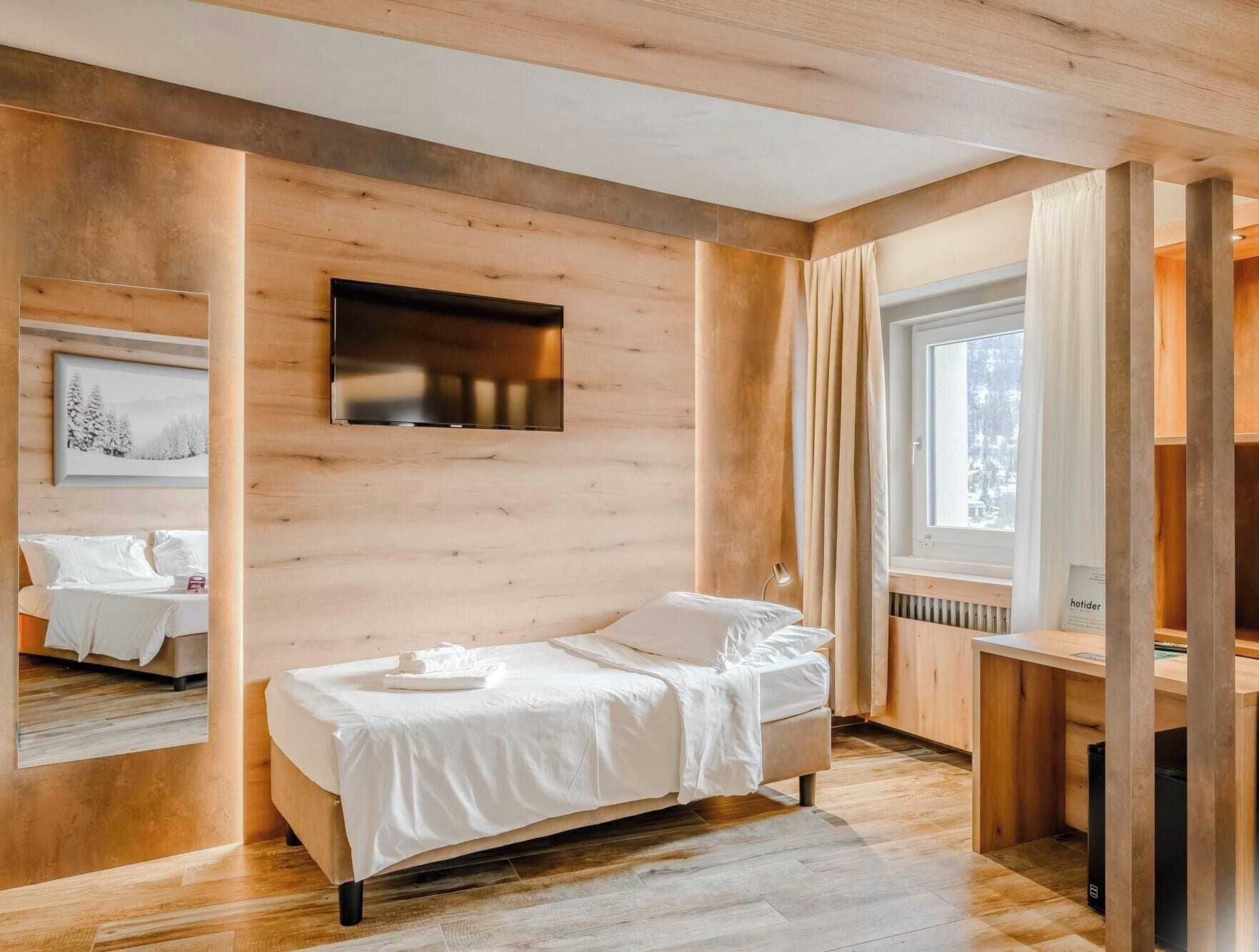 A modern bedroom with a wooden floor, a bed with white linens, a mounted TV, and a window with curtains.