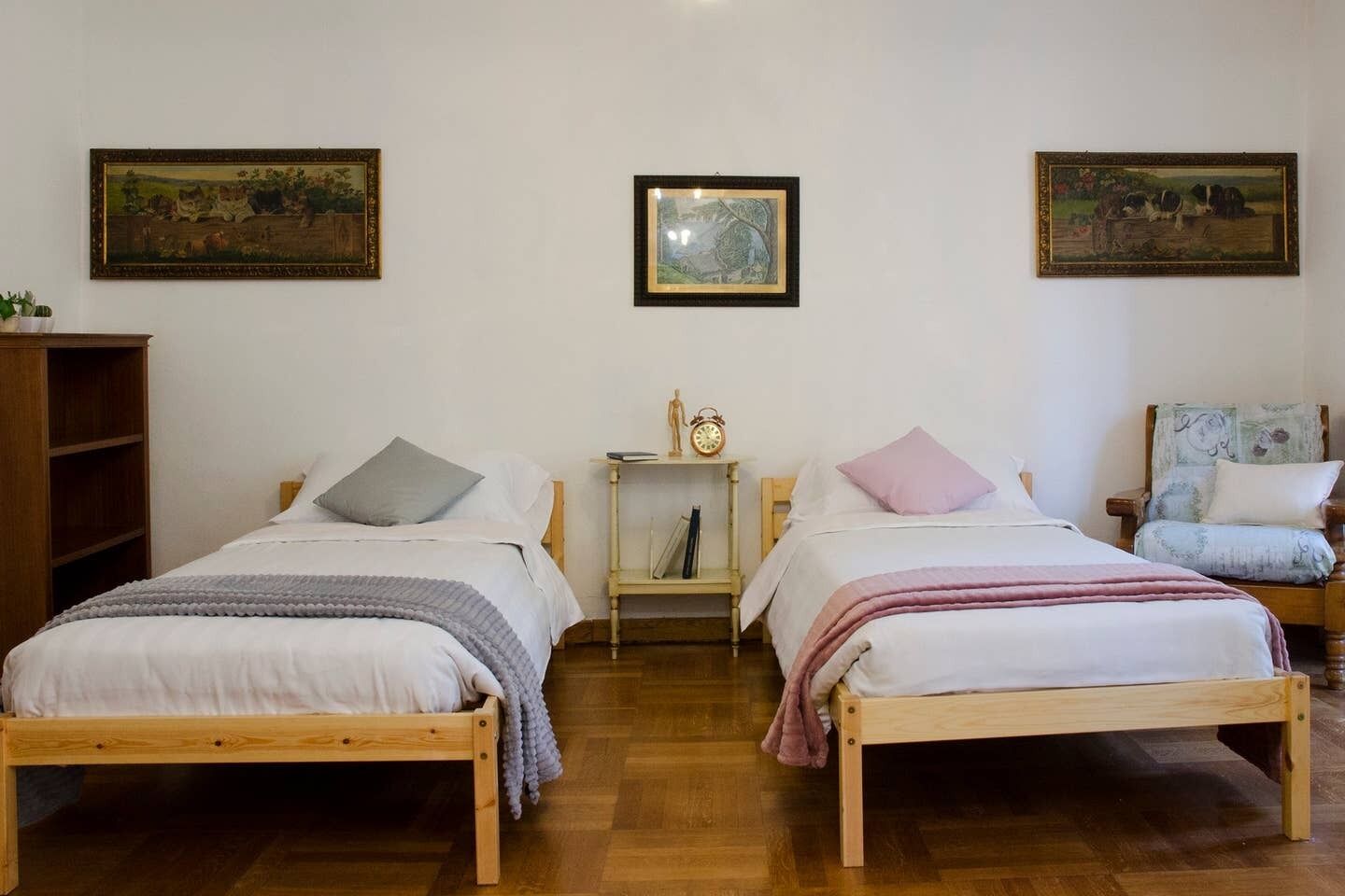 A room with two wooden beds, a small table with a clock, and two framed paintings on the wall.