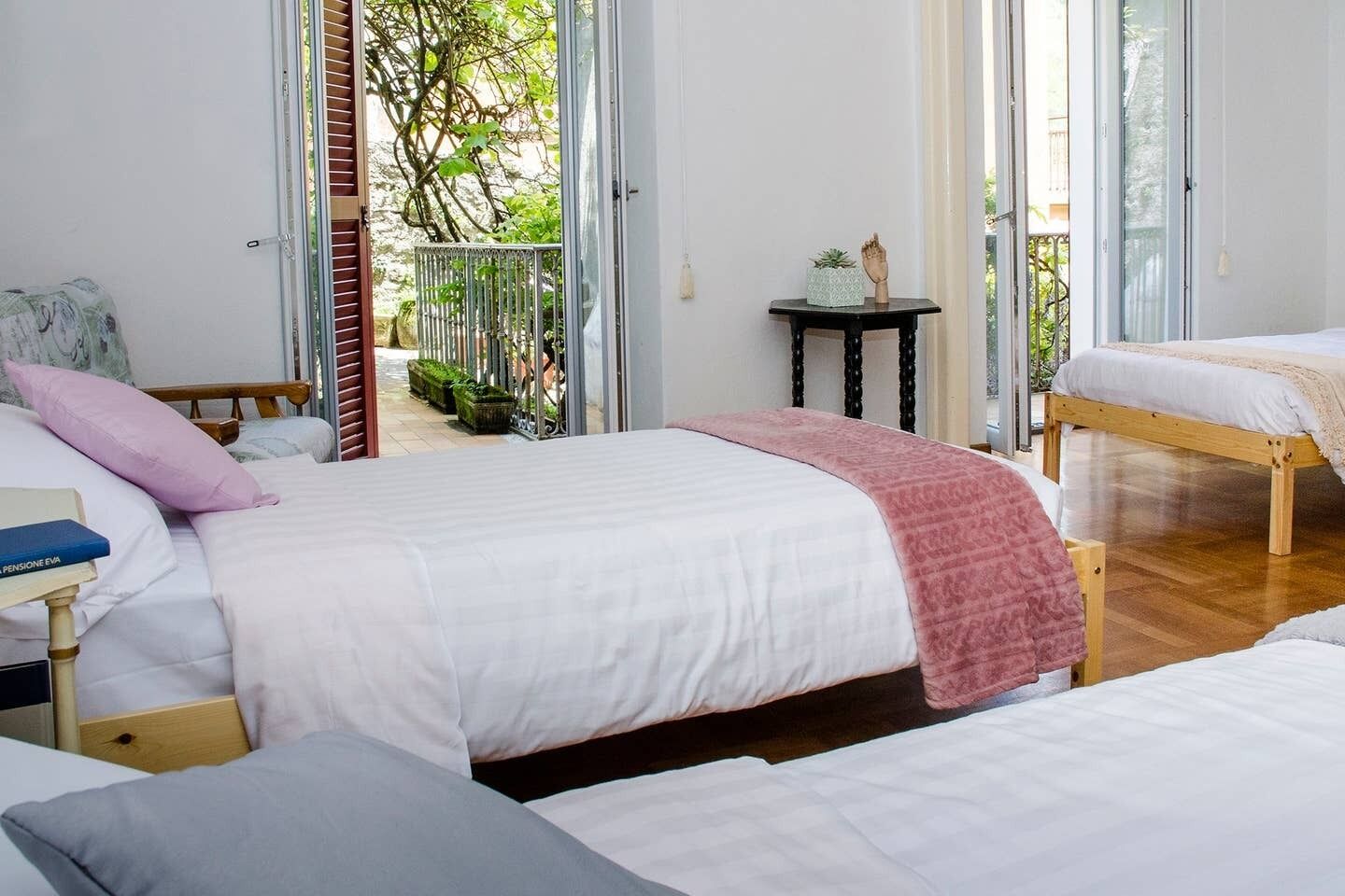 A bedroom with a large bed, a smaller bed, and a nightstand with a book. There is a window with a view of greenery outside.