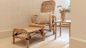 Un fauteuil de relaxation en osier, agrémenté d’un coussin à rayures et d’une petite table sur laquelle est posé un vase rempli de verdure.