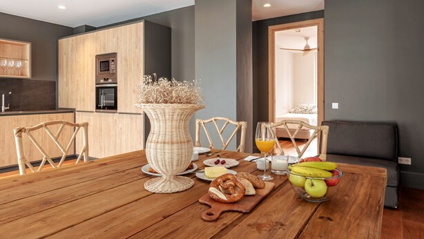 Une cuisine moderne avec des meubles en bois, une table à manger avec des chaises et un coin petit-déjeuner comprenant des fruits, du pain et un vase.