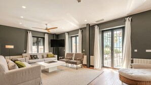 Un salon moderne comprenant un canapé, une table basse et un fauteuil. Un téléviseur à écran plat est fixé au mur, et la pièce est dotée de grandes fenêtres avec des rideaux.