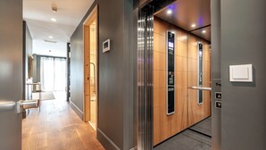 Un couloir moderne avec un plancher en bois, une porte d’ascenseur en verre et un panneau de commande.