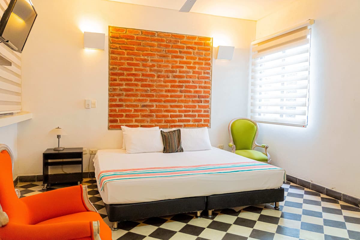 A hotel room with a checkered floor, a bed with a striped bedspread, a green armchair, a brick wall, and a window with blinds.