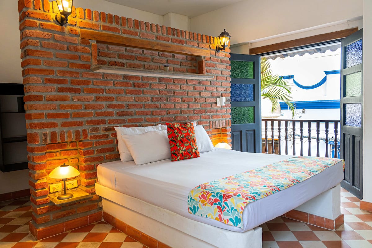 A bedroom with a brick wall, a bed with white bedding, a colorful throw, and a balcony with a view.