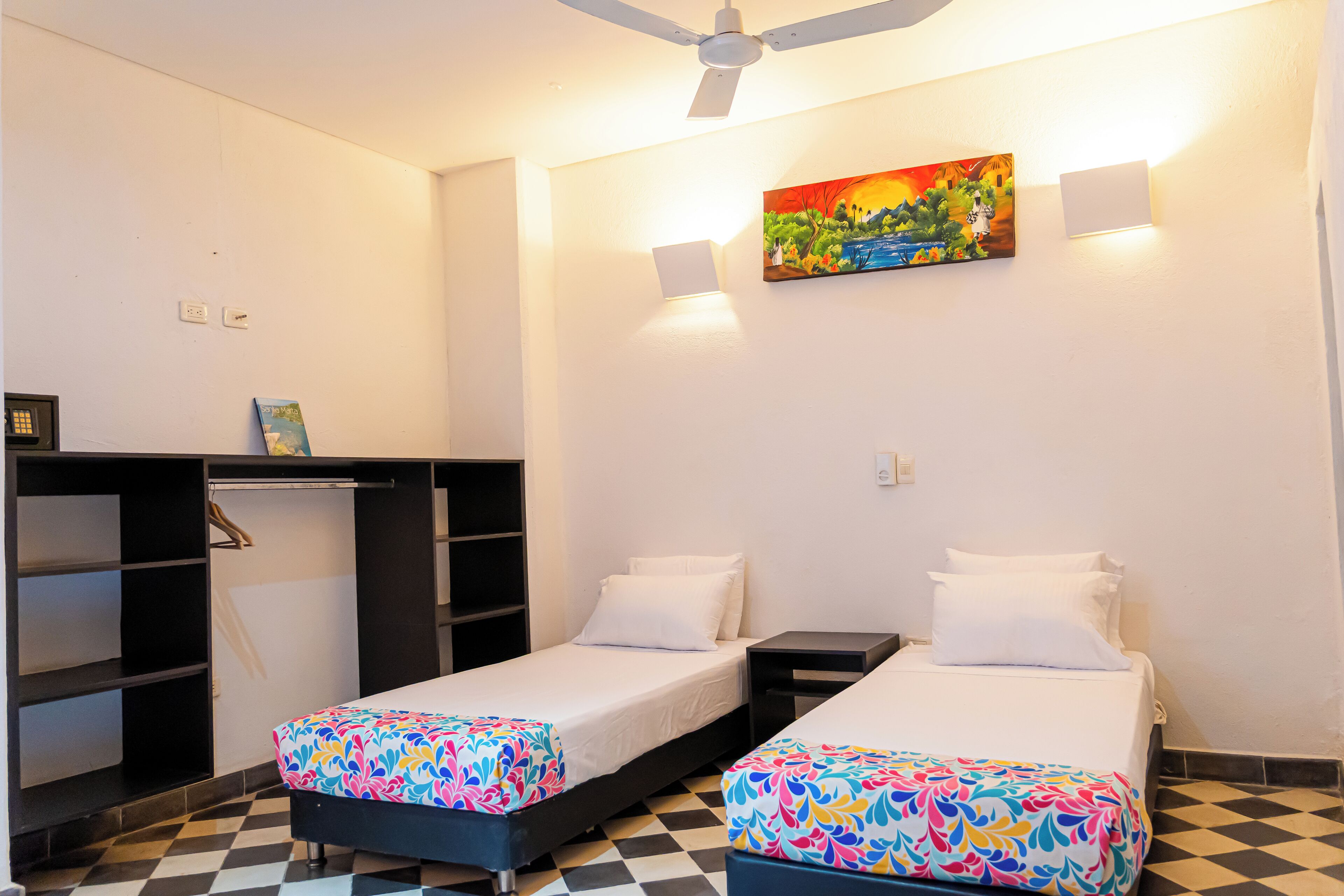 A room with two beds, a desk, and a chair. There is a painting on the wall and a ceiling fan.