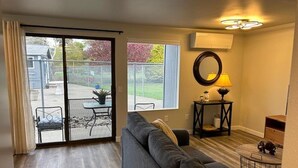 Una sala de estar con un sofá gris, una mesita redonda con una lámpara, y un ventanal corredero que da a un espacio exterior con una mesa y sillas de patio.
