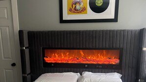 Une chambre à coucher avec un lit, du linge de lit blanc, un disque encadré accroché au mur et une cheminée dans laquelle on peut voir le feu.
