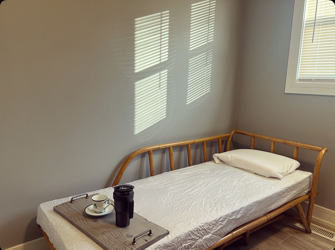 A small, single-bed room with a wooden frame, a white mattress, and a bedside table with a cup and a thermos.