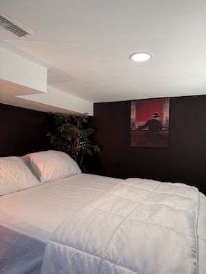 A bedroom with a bed, a ceiling vent, a wall-mounted picture, and a plant.