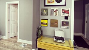 Un salon moderne avec un plancher en bois, un canapé jaune et un banc blanc. Des tableaux encadrés ornent le mur, dont un disque vinyle et une photographie d’oiseau.