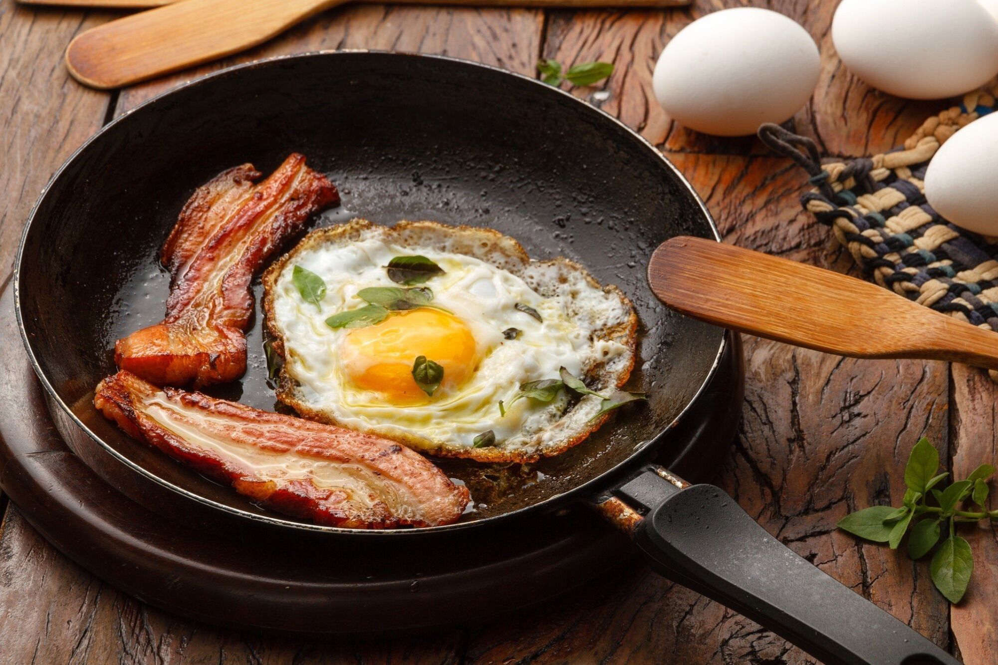 A skillet with bacon and a sunny-side-up egg on a wooden surface.