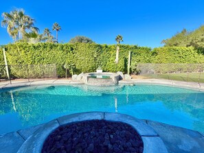 Una piscina con un elemento decorativo de piedra, rodeada por una valla y abundante vegetación.