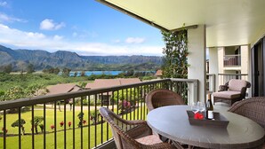 A balcony with wicker furniture, a table with a bottle and glasses, and a view of mountains and a lake. - Hanalei Bay Resort-A/C, Beach, Bali Hai Views from 3 Lanai's, Washer & Dryer (Princeville)