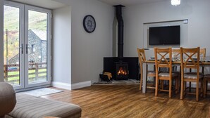 A living room with a fireplace, a dining area with wooden chairs, a television mounted on the wall, and a large window overlooking a stone building.