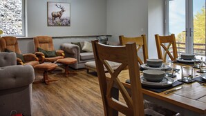 A dining area with a wooden table set for four, surrounded by wooden chairs. A sofa and armchairs are positioned in the background, and a framed deer picture hangs on the wall.